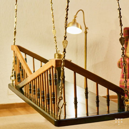 A solid wood swing with a backrest of multiple sleek turned spindles tinted in brown and green, with decorative brass chains