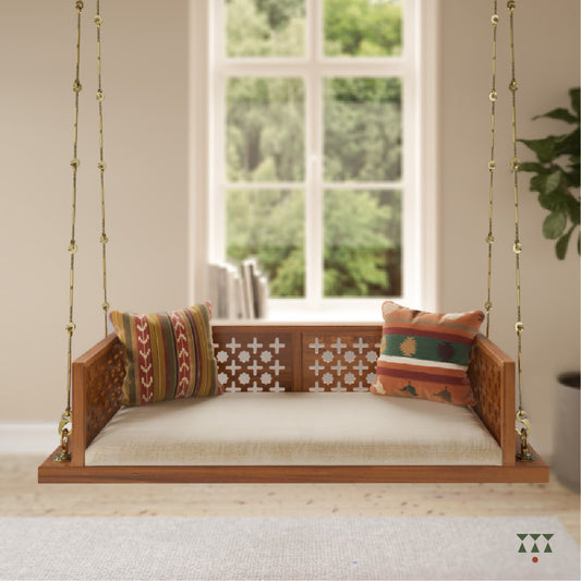 A wooden swing with cutwork patterns, tinted in almond brown with a decorative brass chain, and a fabric seat with cushions