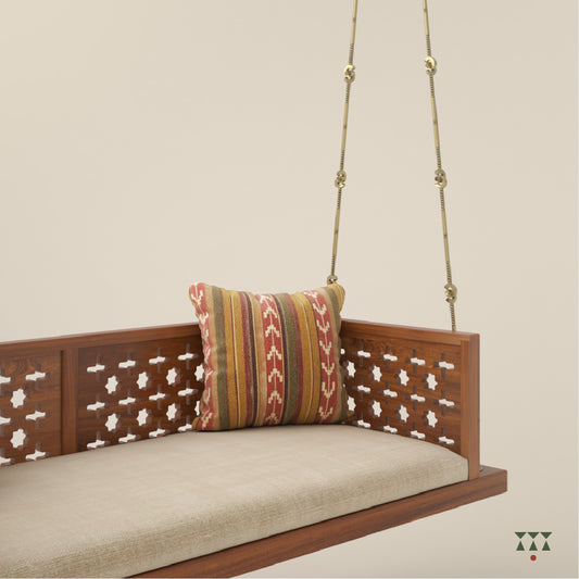 A wooden swing with cutwork patterns, tinted in almond brown with a decorative brass chain, and a fabric seat with cushions