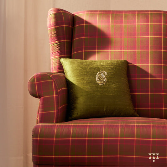 A solid wood wing chair draped in luxurious magenta and green checked silk, and a cushion embroidered with a paisley motif