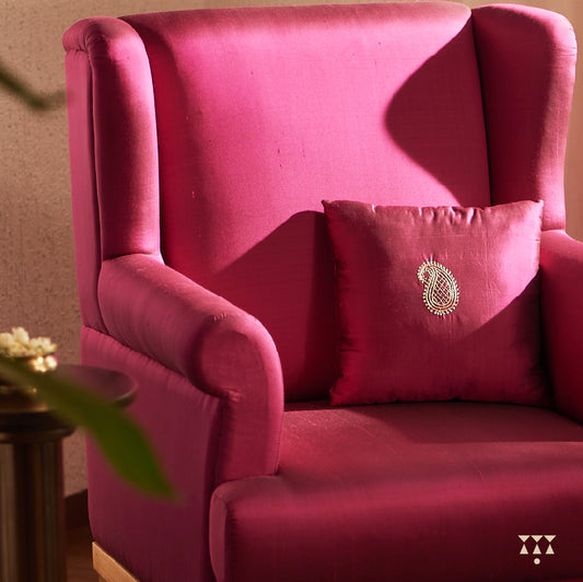 A solid wood wing chair draped in luxurious magenta silk and an embroidered cushion with a paisley motif