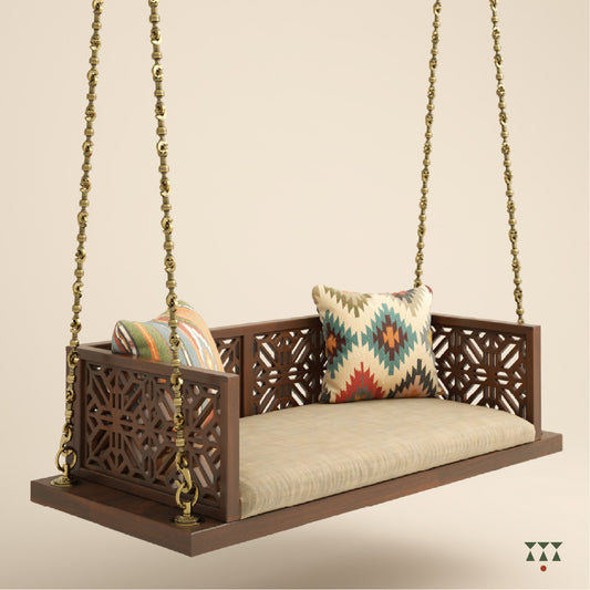 A handcrafted wooden swing with cutwork, tinted in tamarind brown with a decorative brass chain, fabric seat and cushions