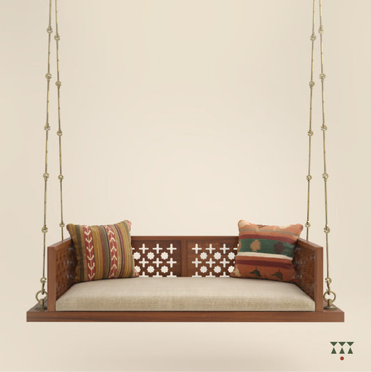 A wooden swing with cutwork patterns, tinted in almond brown with a decorative brass chain, and a fabric seat with cushions