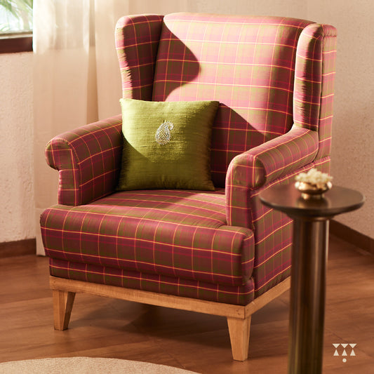 A solid wood wing chair draped in luxurious magenta and green checked silk, and a cushion embroidered with a paisley motif