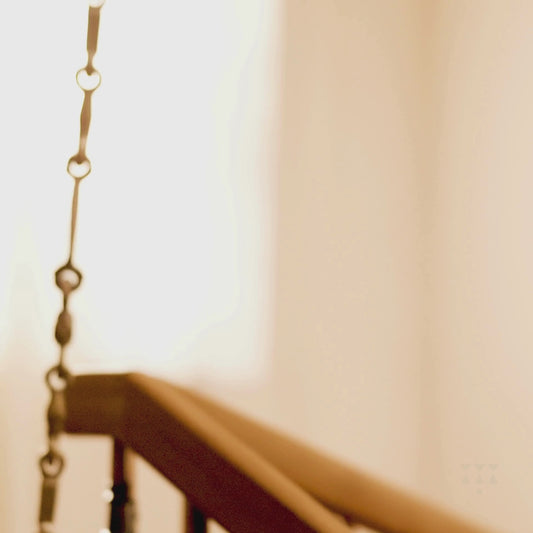 A solid wood swing with a backrest of multiple sleek turned spindles tinted in brown and green, with decorative brass chains